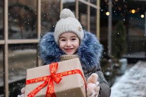 retrato de menina alegre com uma caixa de presente para o natal em uma rua da cidade no inverno com neve em um mercado festivo com enfeites e luzes. agasalhos, gorro de malha, cachecol e peles. espaço de cópia foto