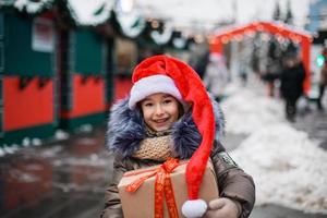 retrato de menina alegre no chapéu de Papai Noel com caixa de presente para o natal na rua da cidade no inverno com neve no mercado festivo com enfeites e luzes de fadas. roupas quentes, cachecol de malha e peles. ano Novo
