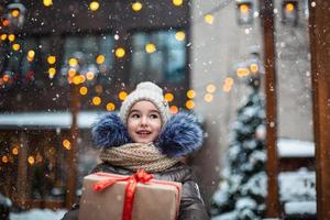 retrato de menina alegre com uma caixa de presente para o natal em uma rua da cidade no inverno com neve em um mercado festivo com enfeites e luzes de fadas. agasalhos, gorro de malha, cachecol e peles. ano Novo foto