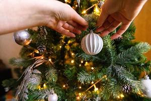 mãos de mulher decorando a árvore de natal. árvore de natal com enfeites de wite e prata, enfeites de brinquedo e bola. design de interiores de estilo clássico moderno. véspera de natal em casa, tempo de celebração. foto