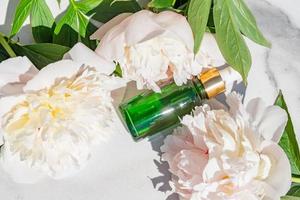 frasco de soro cosmético com óleo essencial e flores de peônia em fundo de mármore branco. cosméticos naturais, conceito de cuidados com a pele. foto