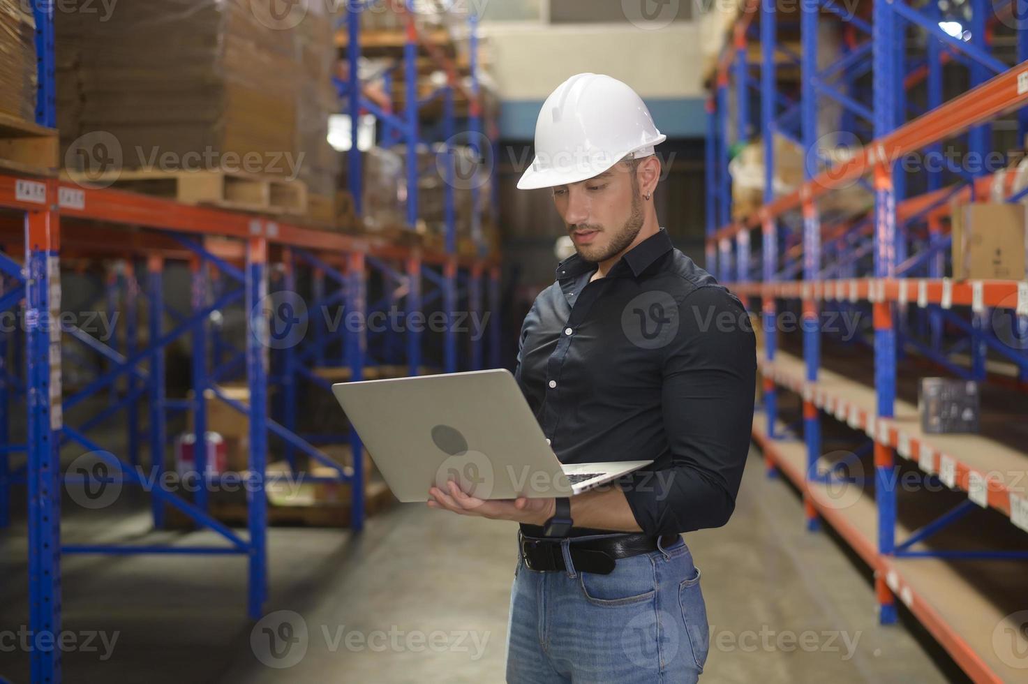 junger kaukasischer männlicher arbeiter, der helm mit laptop in modernem lager trägt. foto