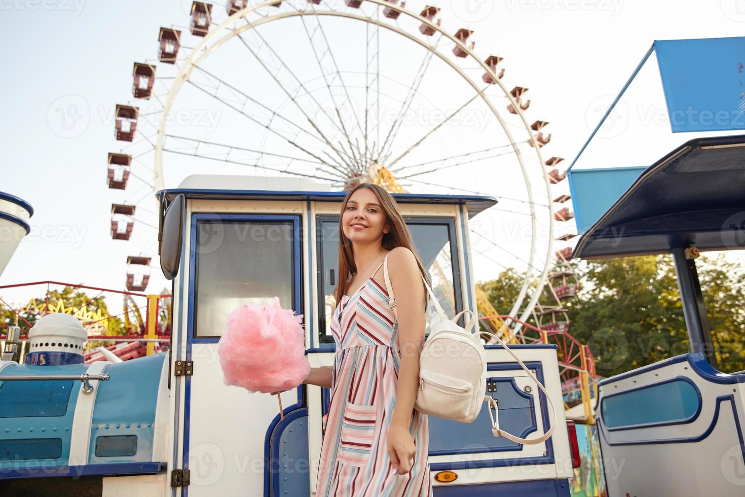 charmante junge brünette dame mit langen braunen haaren, die an einem sonnigen warmen tag über dem riesenrad posiert, ein romantisches kleid und einen weißen rucksack trägt und zuckerwatte am stab hält foto