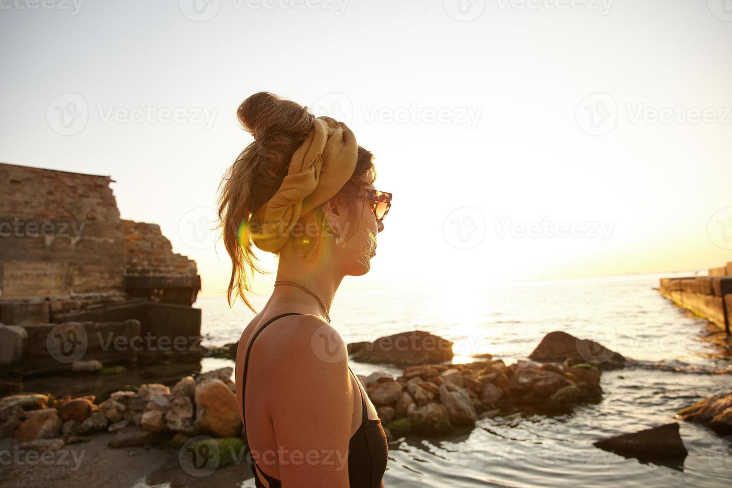 Outdoor-Foto einer jungen dunkelhaarigen Frau mit lässiger Frisur, die am frühen Morgen mit Blick auf das Meer posiert und einen schwarzen Badeanzug, ein Stirnband und eine Sonnenbrille trägt foto