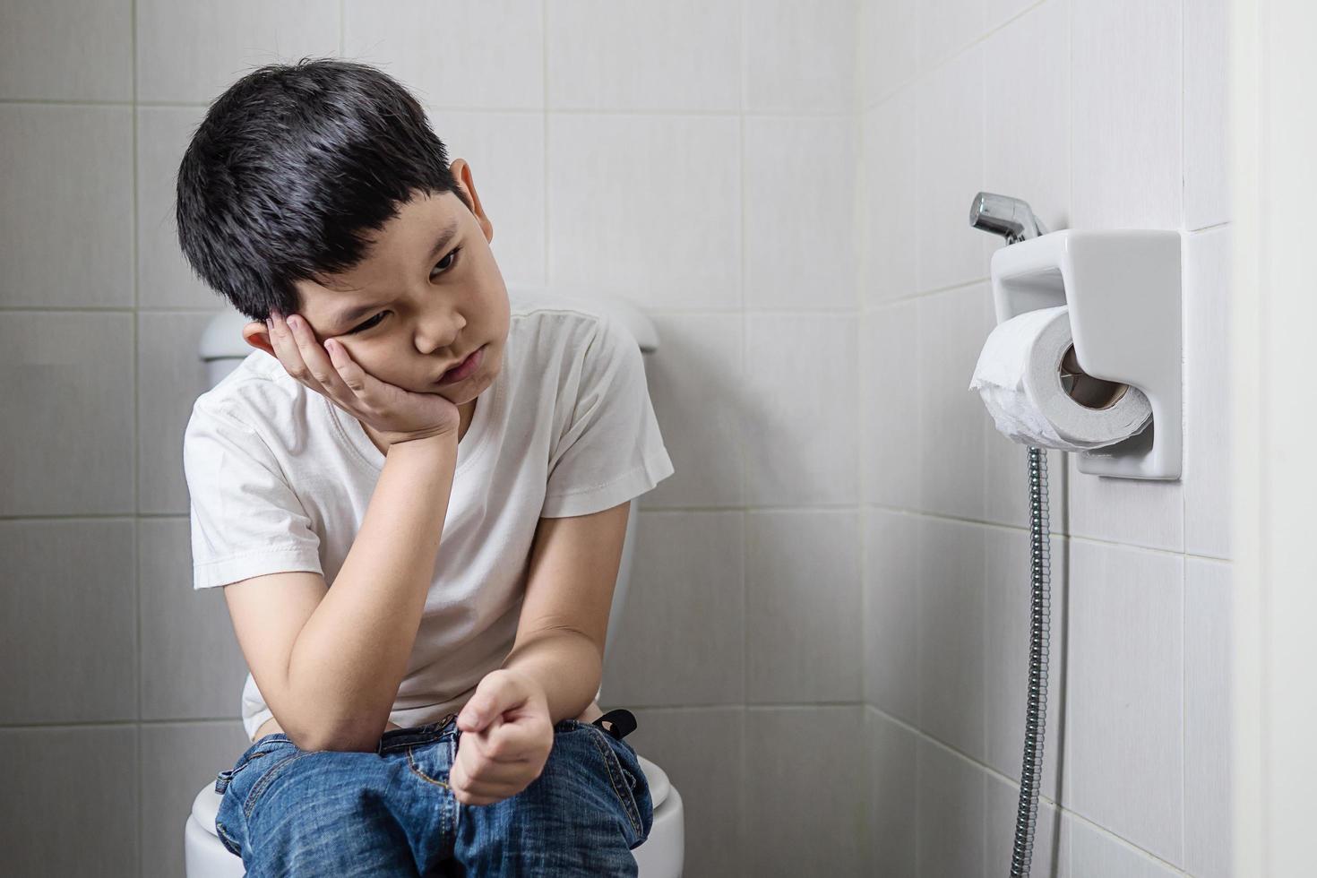 asiatischer junge, der auf toilettenschüssel sitzt und seidenpapier hält - gesundheitsproblemkonzept foto