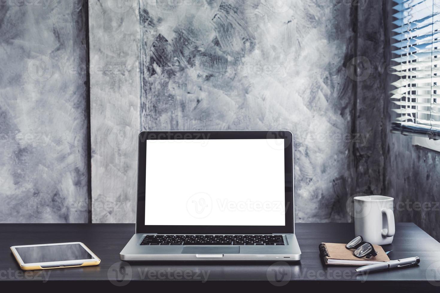 Laptop mit weißem, leerem Bildschirm, weißer Tasse Kaffee, Tablet, Notizbuch, Stift und Brille auf dem Schreibtisch foto
