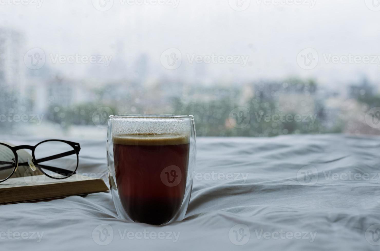 ein glas heißen kaffee mit buch und brille morgens auf dem bett mit regentropfen auf dem fenster. zu hause bleiben und entspannendes konzept. foto
