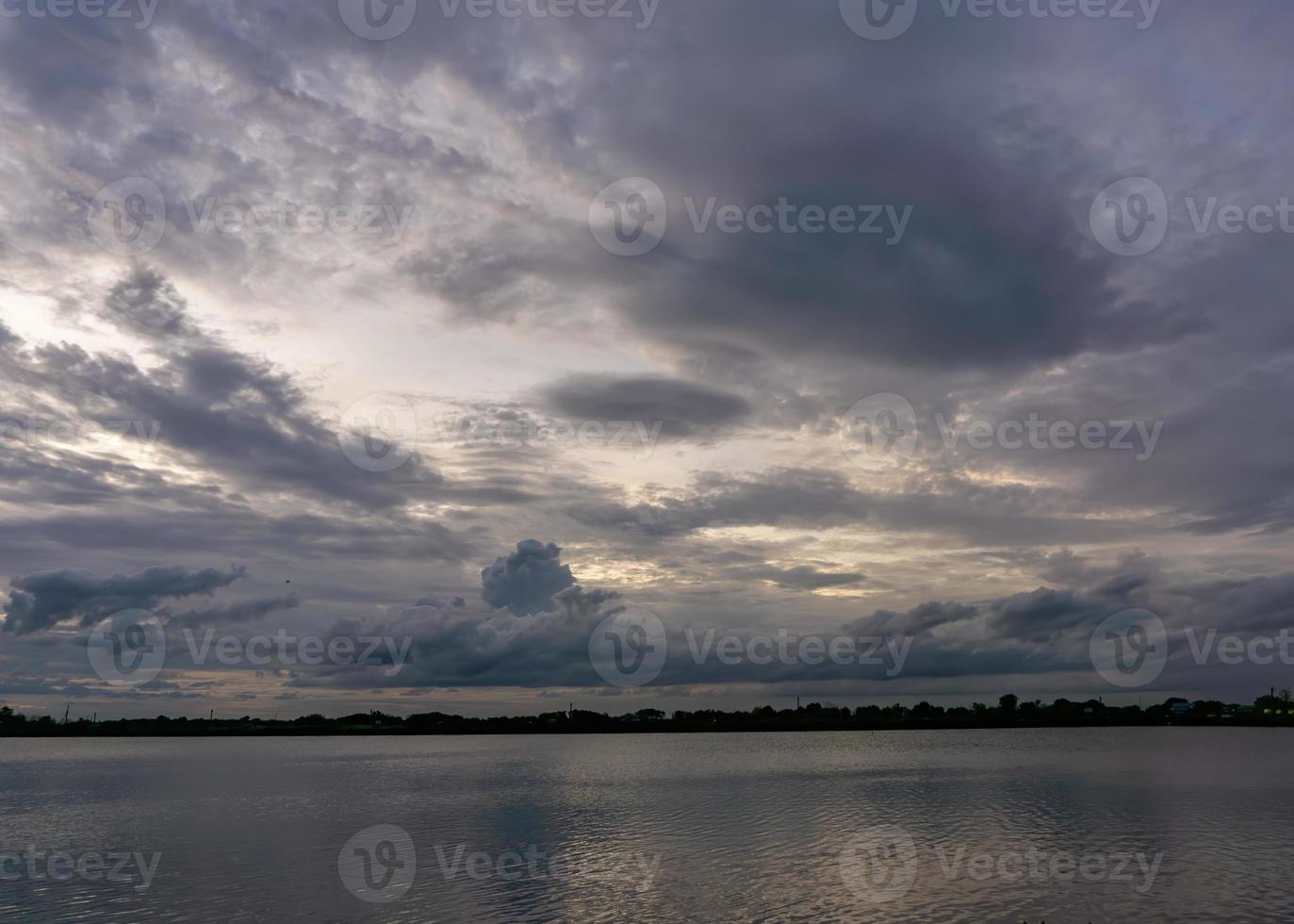 Die Wolken bedecken den Himmel und verdunkeln das Licht der Sonne bei Sonnenuntergang. Die Skyline teilt den Teich und der Himmel ist mit Bäumen bedeckt. foto