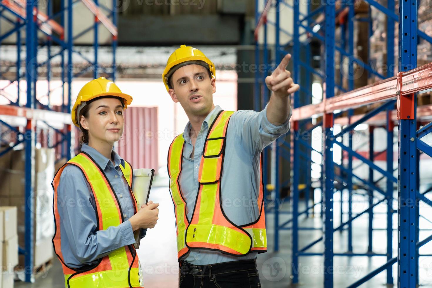 Ingenieur, der eine Sicherheitsweste trägt, die die Maschinenarbeit steuert, spricht mit einem Hilfsingenieur, der zuerst die Sicherheit für Arbeiter überprüft. Kontrollkästchen Sicherheitsbeauftragter im Lager. foto