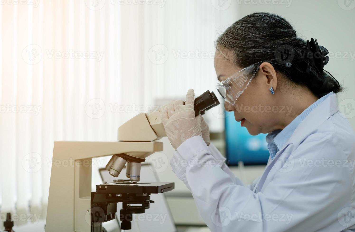 medizinisches entwicklungslabor wissenschaftlerin, die unter mikroskop schaut, analysiert petrischalenprobe. im hintergrund großes pharmazeutisches labor mit spezialisten für medizin, biotechnologieforschung foto