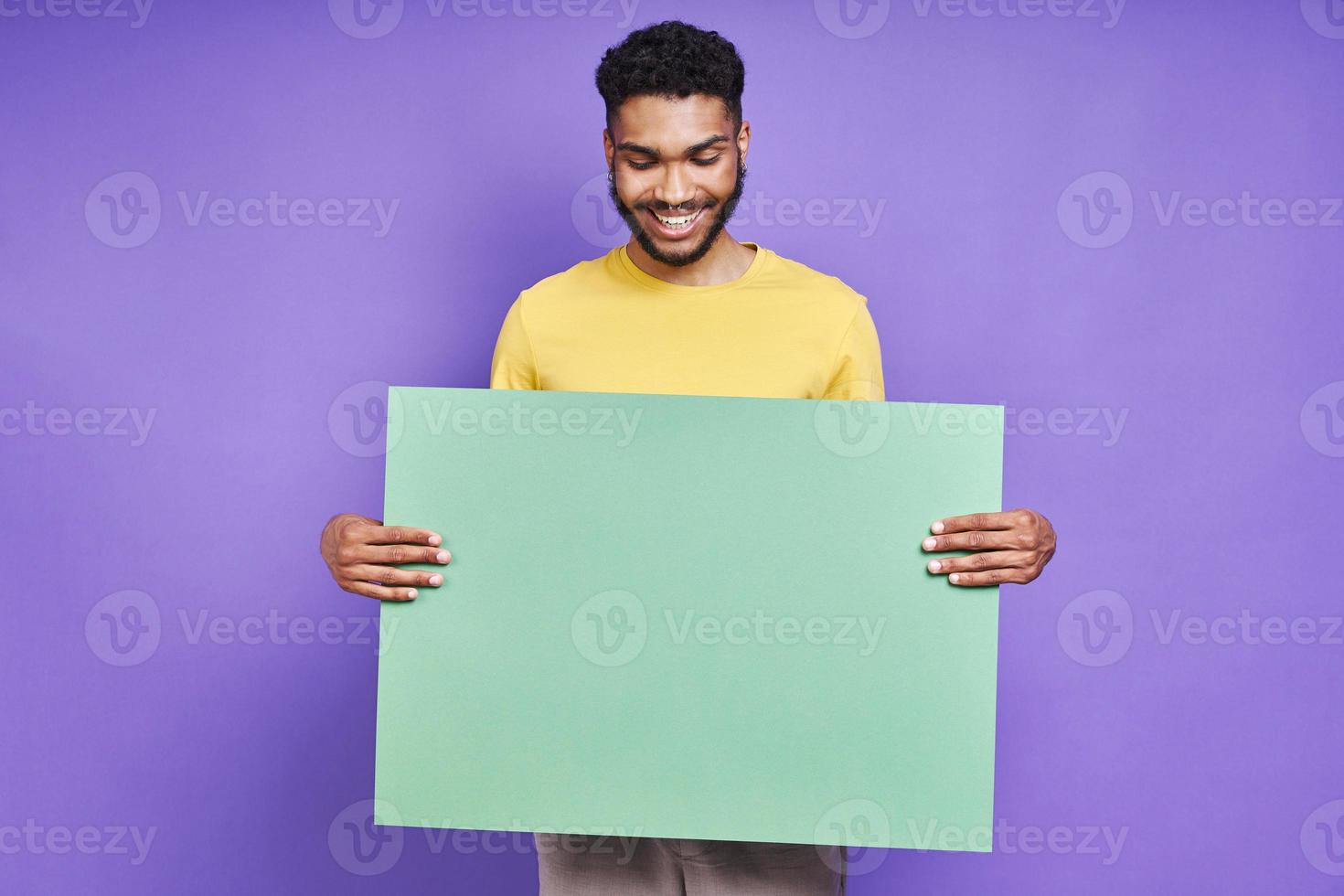 hübscher afrikanischer mann, der grünes banner hält und lächelt, während er vor lila hintergrund steht foto