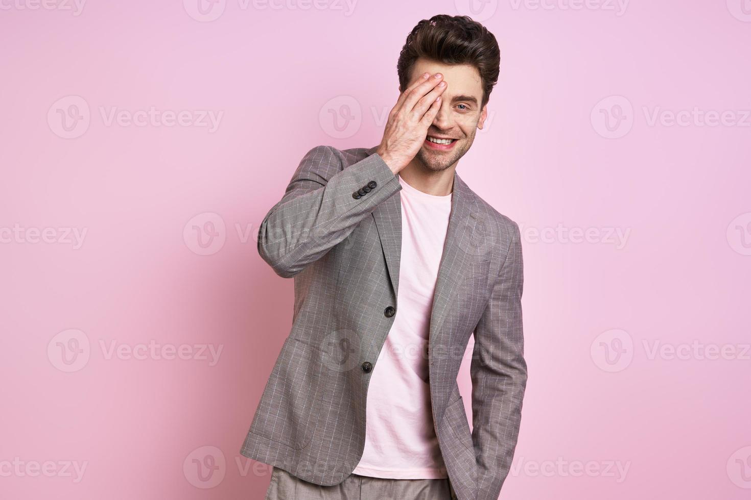 hübscher junger Mann im Anzug, der das halbe Gesicht mit der Hand bedeckt, während er vor rosa Hintergrund steht foto