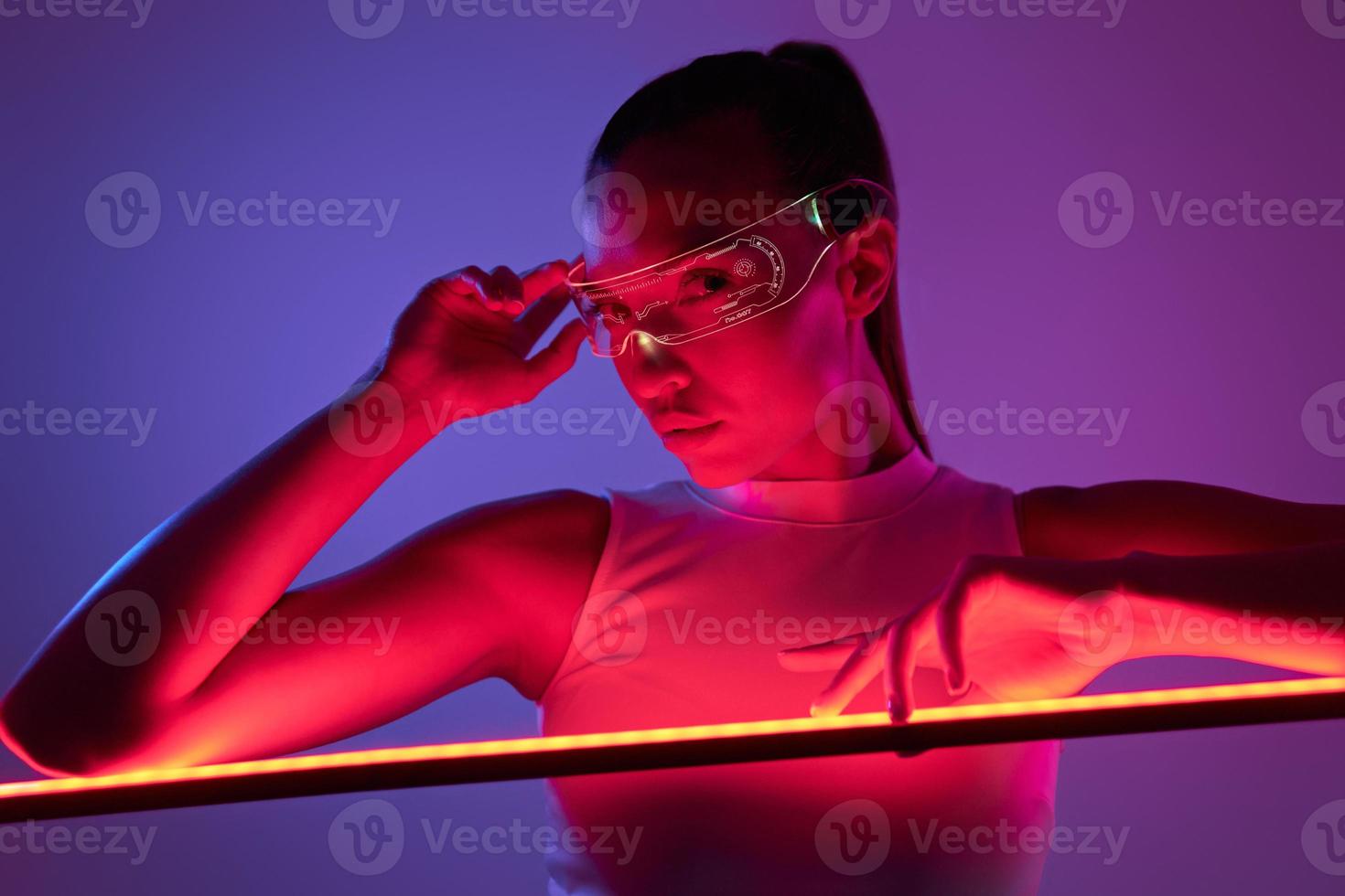 Schöne Frau mit futuristischer Brille, die sich vor dunklem Hintergrund an die LED-Lampe lehnt foto