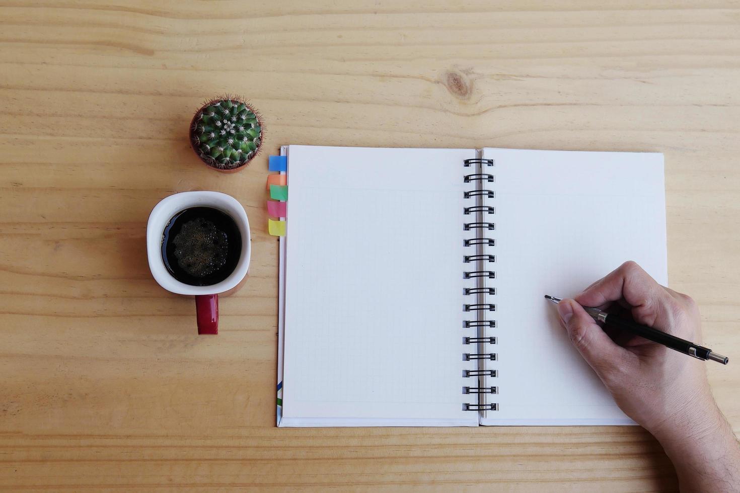 Draufsicht des Mannes, der Notizbuch auf Holztisch mit Kaffeetasse und kleinem Kaktustopf schreibt foto