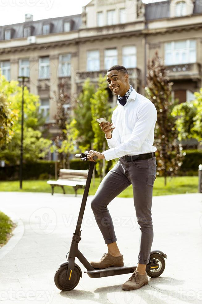 junger Afroamerikaner mit Handy beim Stehen mit Elektroroller auf einer Straße foto