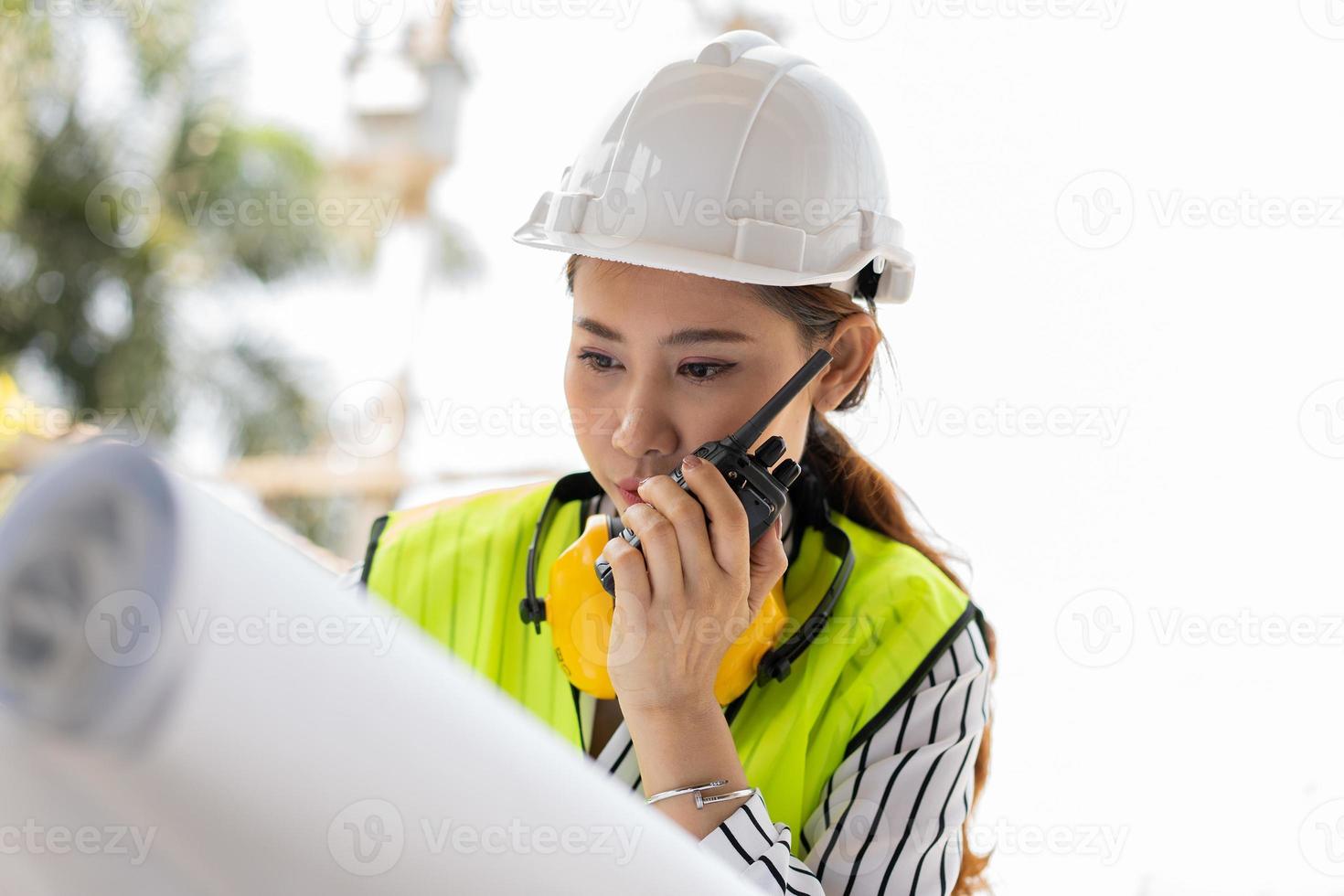 asiatische ingenieure oder junge architekten setzen aus sicherheitsgründen einen helm auf und sehen sich den bauplan an, um die baustelle der gebäudefabrik zu inspizieren, und verwenden das walkie-talkie für ein gespräch mit einem auftragnehmer. foto