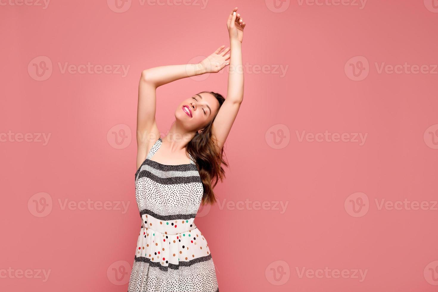 Attraktive, stilvolle junge Frau mit romantischem Lächeln und geschlossenen Augen, die Hände hochhalten und während des Fotoshootings auf rosafarbenem Hintergrund in die Kamera posieren foto