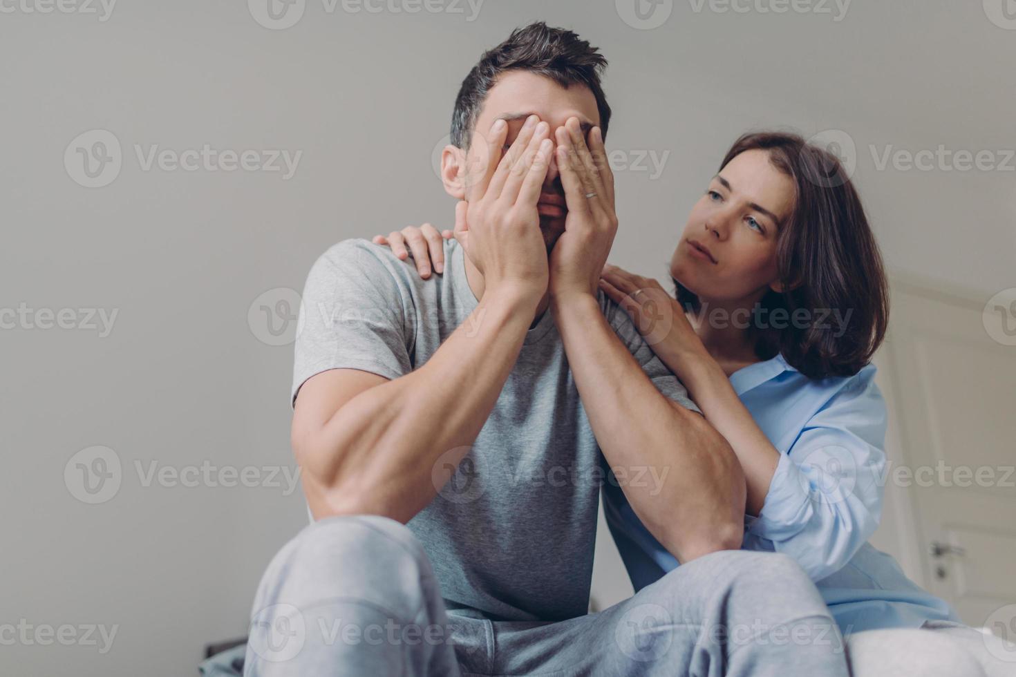 Foto eines depressiven Mannes, der lässig gekleidet ist, seine schöne Frau tröstet ihn, sitzt zusammen im Schlafzimmer, hat Probleme im Familienleben. liebevolle frau tröstet ehemann in schwierigen situationen.