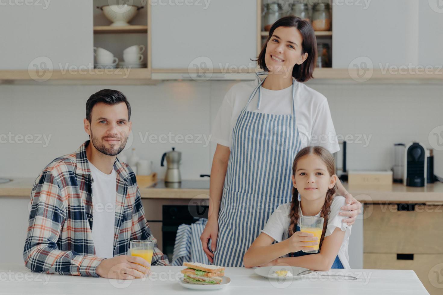 familie besteht aus mutter, vater und tochter posieren zusammen in moderner küche, trinken frischen saft, essen sadwiches, genießen häusliche atmosphäre. Schöne Frau in Schürze umarmt kleines Kind mit Liebe foto