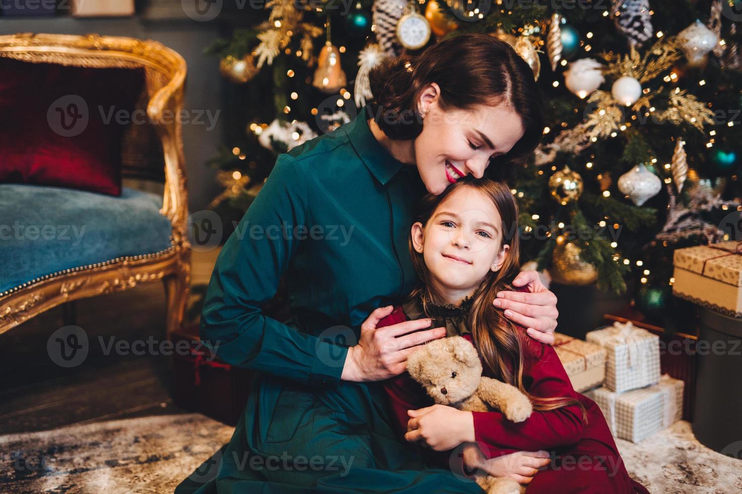 Die liebevolle Mutter umarmt ihre kleine Tochter, während sie neben dem geschmückten Weihnachtsbaum zusammensitzt. Kleines entzückendes Mädchen, das froh ist, ein Neujahrsgeschenk von ihrer Mutter zu erhalten. Beziehungskonzept. foto