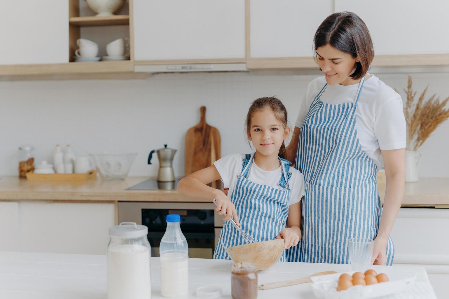 lächelnde mutter und tochter bereiten leckere kekse zu, mädchen verquirlt zutaten in schüssel mit schläger, hilft mutter in der küche, bereitet festliches abendessen zu, modernes wohninterieur. Familie, Kochen, Lifestyle-Konzept foto