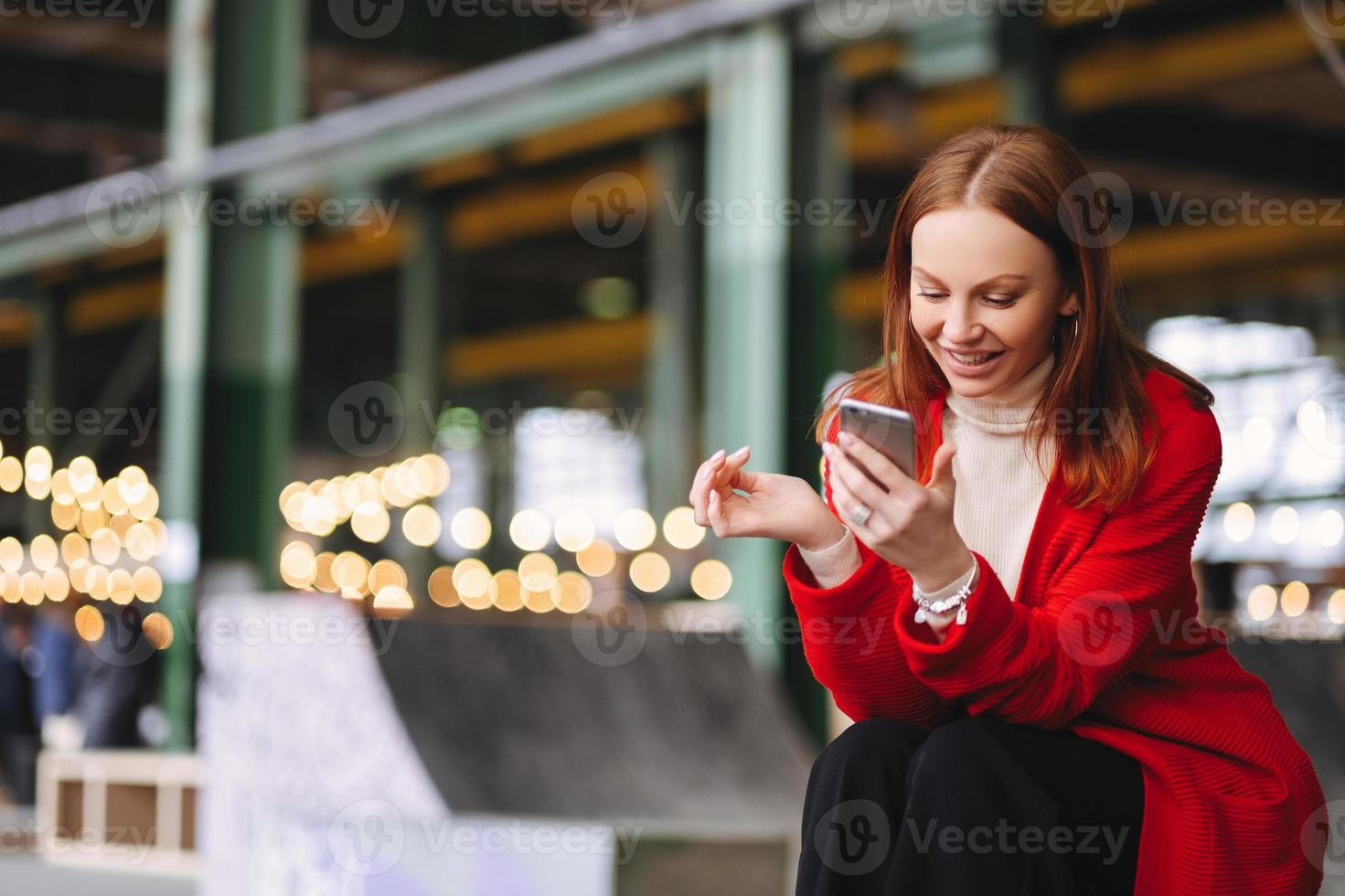 zufriedene junge europäerin im roten mantel, hält handy in der hand, liest benachrichtigung, ist mit dem drahtlosen internet verbunden, hat freizeit, posiert im freien, wartet auf jemanden, genießt die online-kommunikation foto