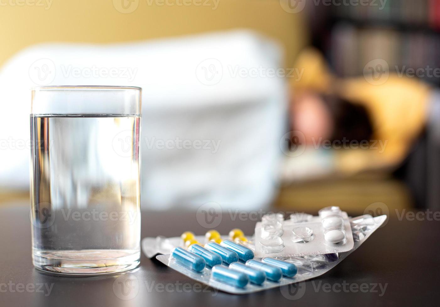 tabletten und ein glas wasser auf dem tisch im hintergrund liegt ein kranker mensch im bett foto