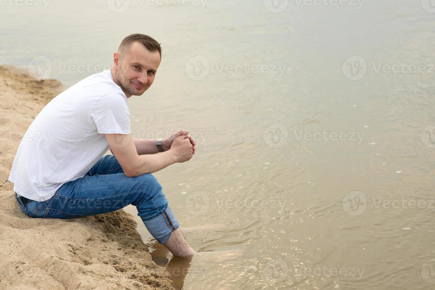 Bild eines einsamen, positiven und lächelnden Mannes, der am Flussufer sitzt und seine Beine ins Wasser hält foto