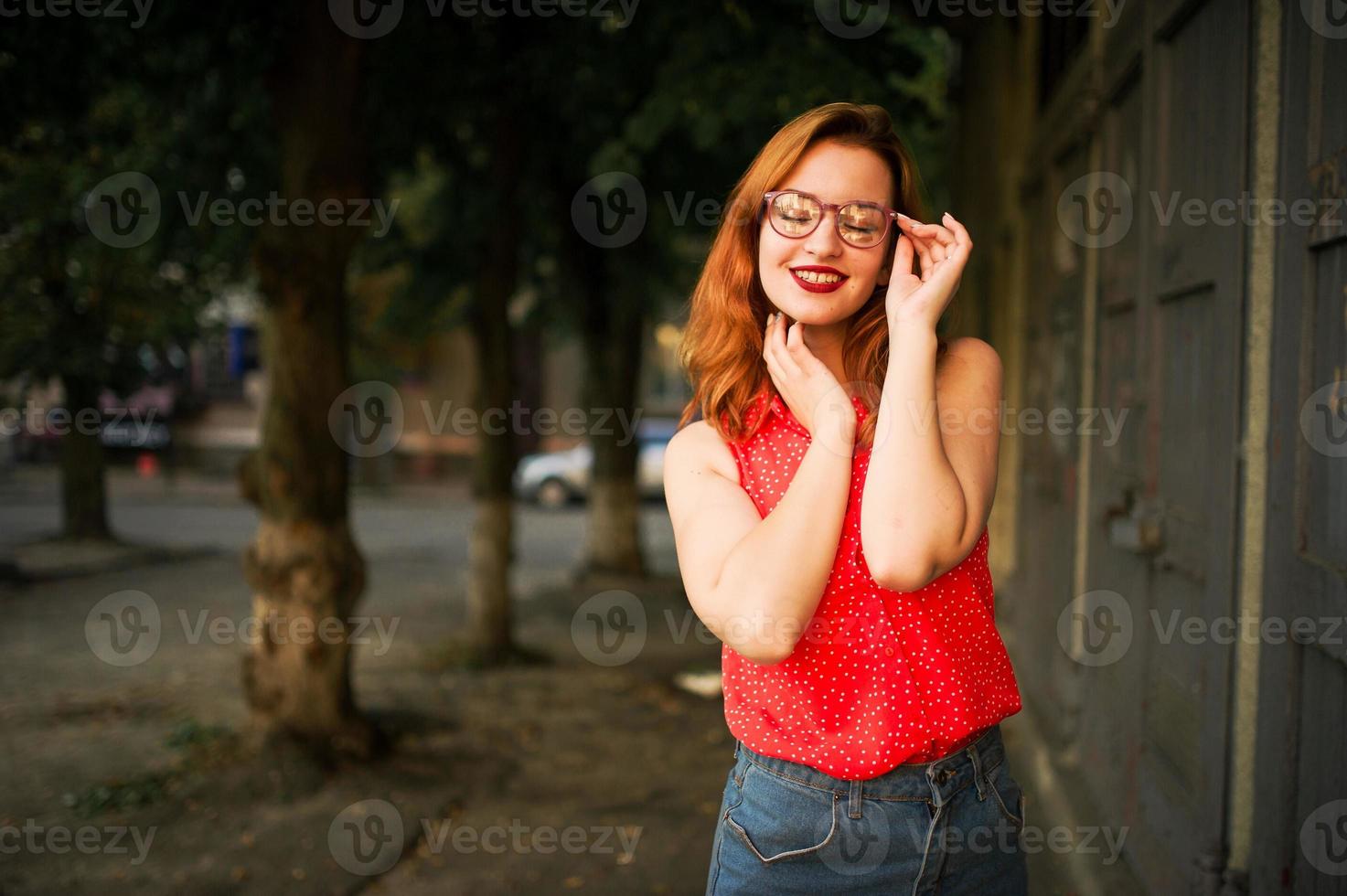attraktive rothaarige frau mit brille, tragen auf roter bluse und jeansrock posieren. foto