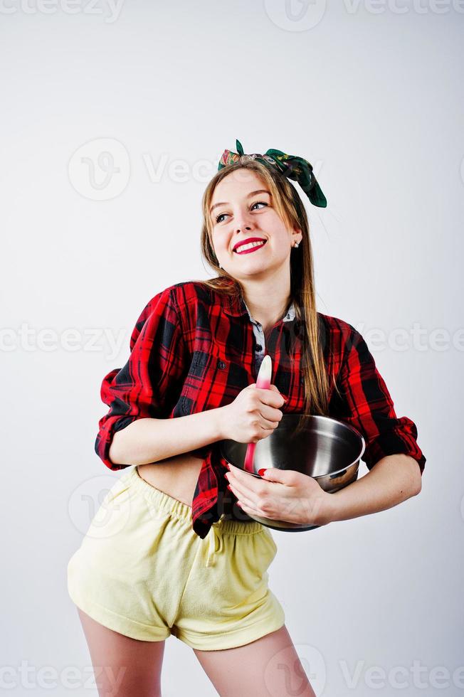 junge lustige Hausfrau in kariertem Hemd und gelben Shorts Pin-up-Stil mit Topf und Küchenlöffel isoliert auf weißem Hintergrund. foto