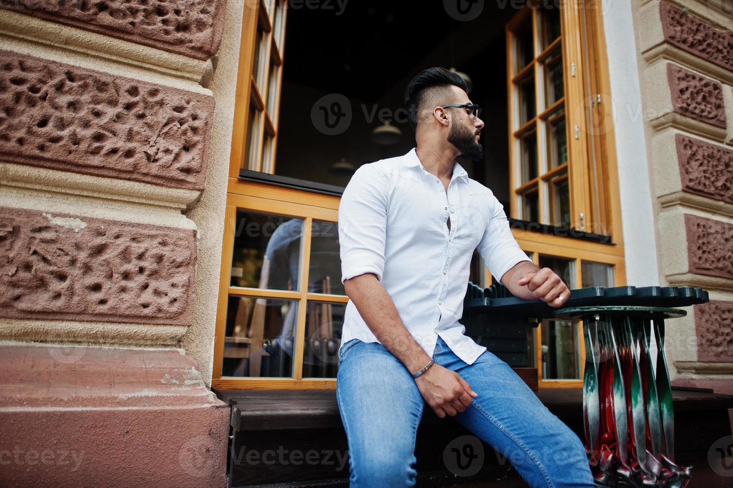 Stilvolles großes arabisches Mannmodell in weißem Hemd, Jeans und Sonnenbrille, das auf der Straße der Stadt sitzt. Bart attraktiver arabischer Typ gegen offenes Fenster des Restaurants im Freien. foto