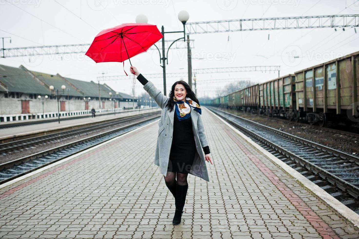 Brünettes Mädchen im grauen Mantel mit rotem Regenschirm im Bahnhof. foto