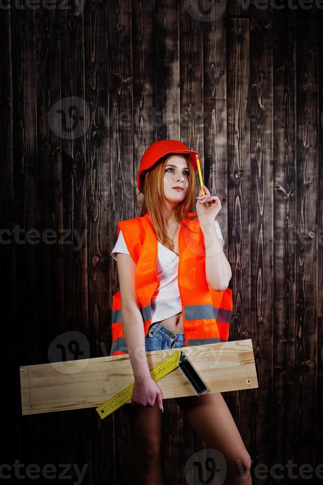 ingenieurfrau in orange schützt helm und baujacke vor hölzernem hintergrund, der brett und lineal hält. foto