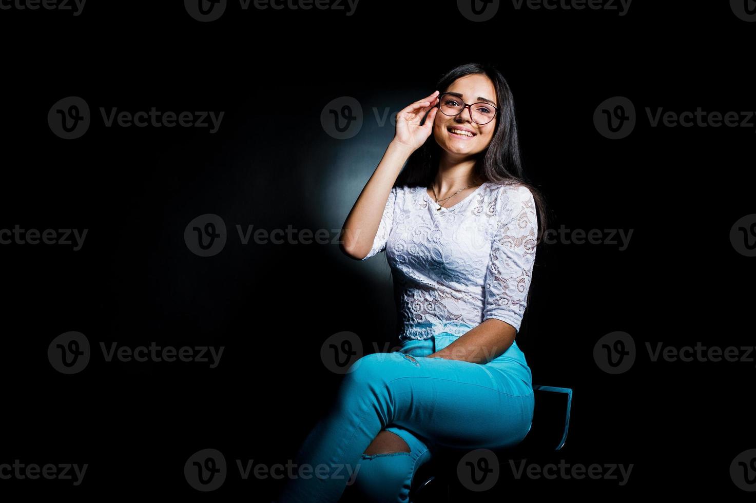 Porträt einer attraktiven jungen Frau in weißem Oberteil und blauer Hose, die mit ihrer Brille im Dunkeln posiert. foto