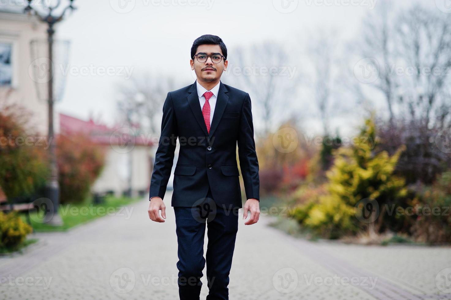 indischer junger mann mit brille, trägt einen schwarzen anzug mit roter krawatte im freien. foto