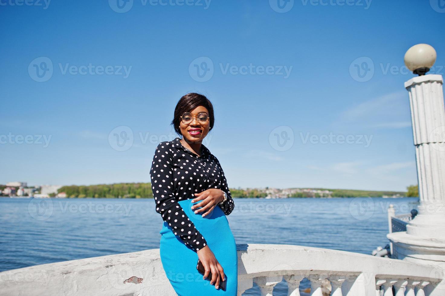 Stilvolles afroamerikanisches Modelmädchen mit Brille, blauem Rock und schwarzer Bluse posierte im Freien auf einer Brücke am See. foto