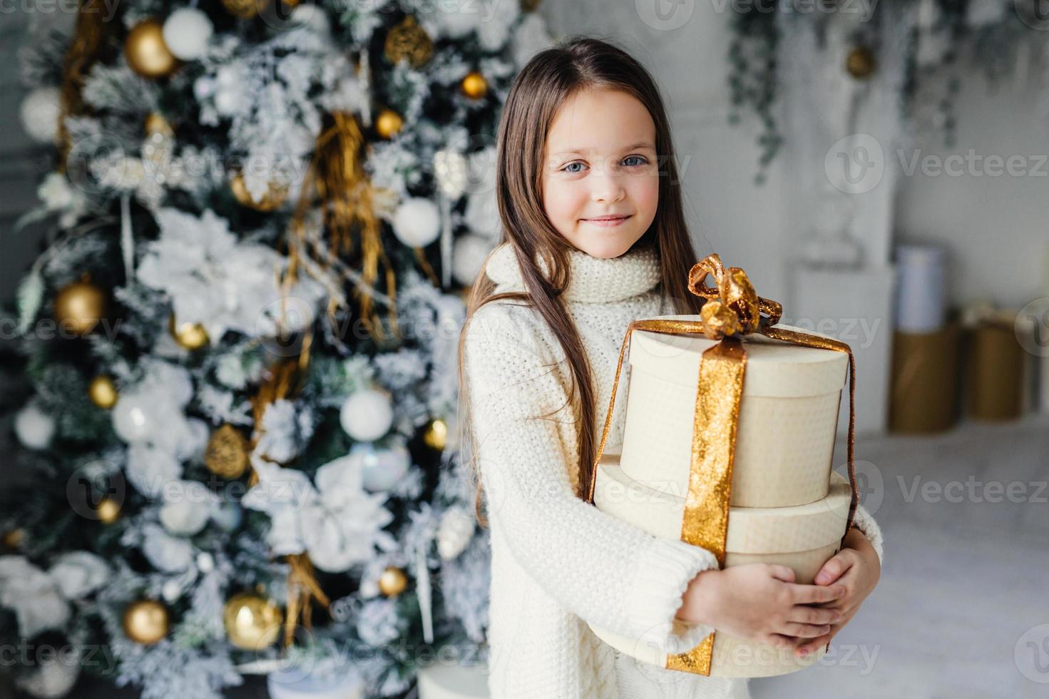 innenaufnahme eines entzückenden niedlichen hübschen kleinen weiblichen kindes trägt warme strickkleidung, hält ein riesiges verpacktes geschenk, erhält vom weihnachtsmann, steht in der nähe eines geschmückten schönen neujahrsbaums foto