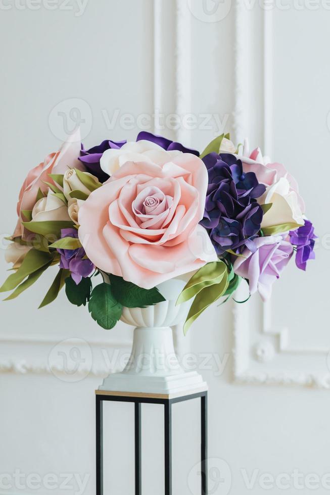 vertikale Aufnahme von künstlichen Blumen auf Blumenständer und hoher Vase vor weißem Hintergrund in einer geräumigen Halle, die als Dekorationsobjekt verwendet wird. foto