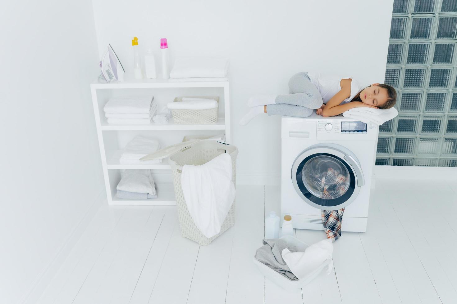 aufnahme eines müden kleinen kindes, das auf der waschmaschine schläft, ein weißes weiches handtuch als kissen verwendet, angenehme träume hat, korb und becken auf weißem boden, sich müde fühlt, nachdem es kleidung in die waschmaschine geladen hat, zu hause ist foto