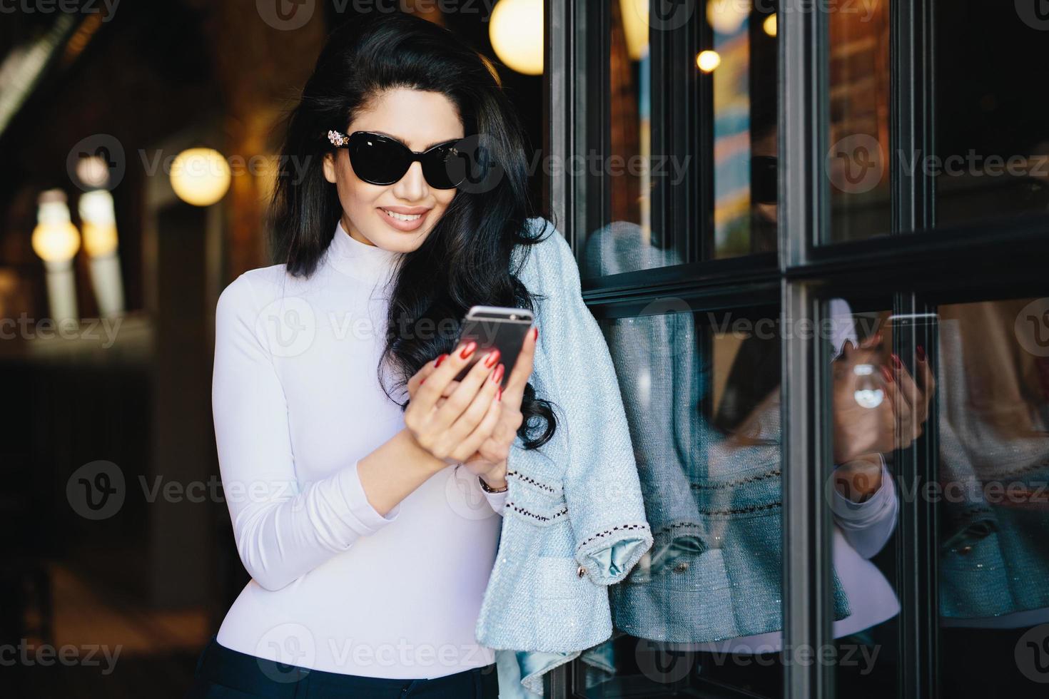 Stilvolle Frau mit dunklem, welligem Haar in weißer Bluse mit Jacke und großer Sonnenbrille mit Smartphone, die online mit ihrem Freund kommuniziert. Brünette Frau, die neue Anwendungen aktualisiert foto