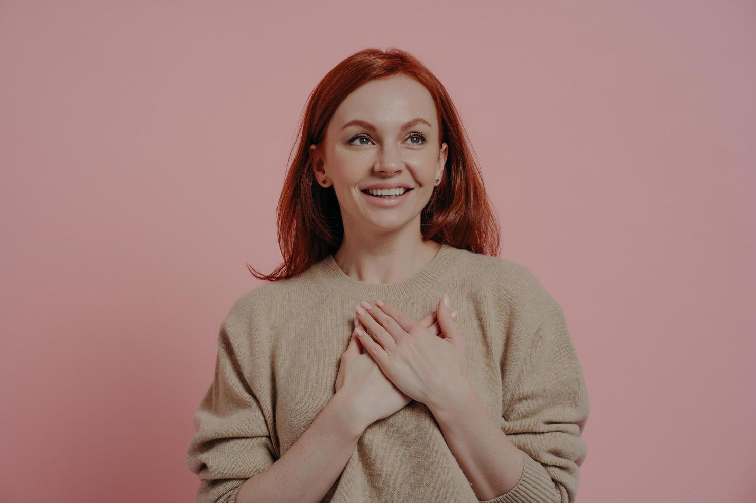 junge glückliche rothaarige Frau, die Hände auf die Brust legt, während sie isoliert auf rosa Hintergrund steht foto