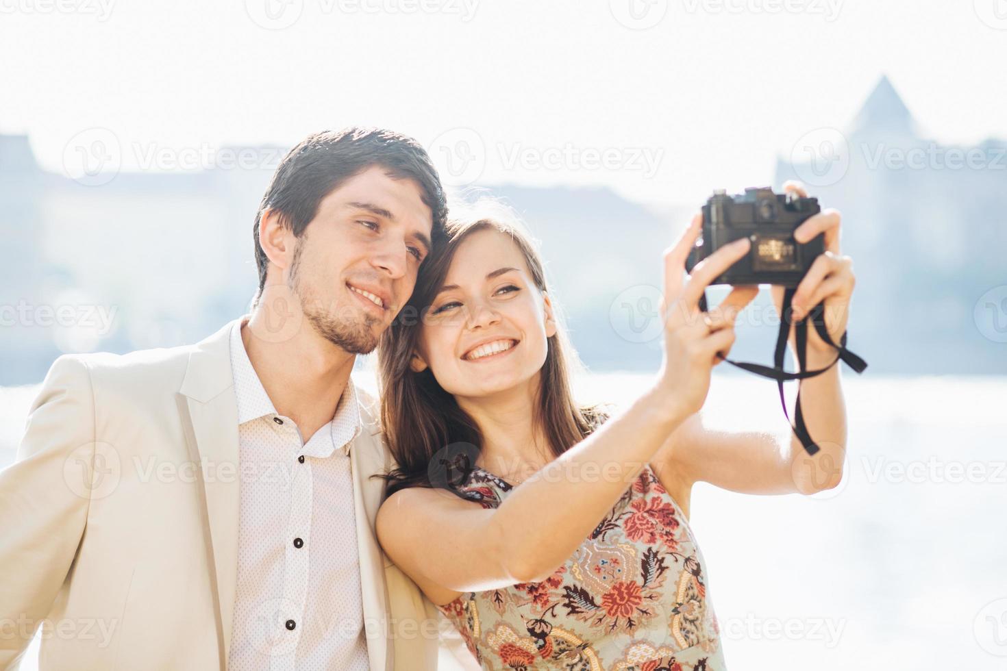 glückliches verliebtes paar posiert für selfie in moderner professioneller kamera, spazieren im freien, erkunden unbekannte orte in großen touristenstädten, modellieren vor wunderbarer landschaft mit wasser im hintergrund foto