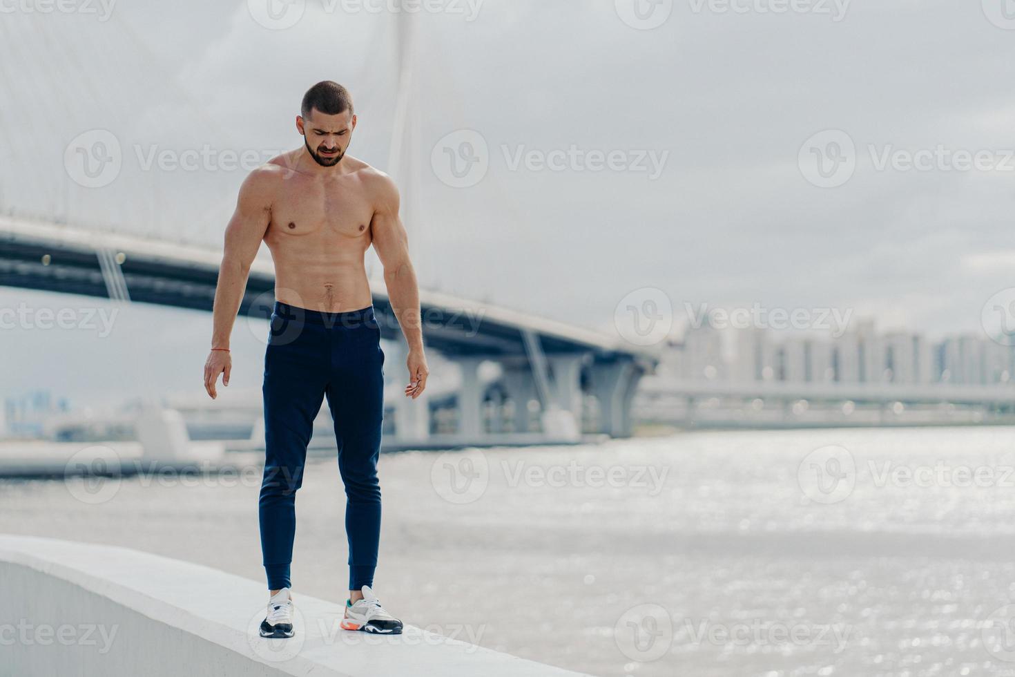 sportlicher bärtiger mann mit muskulösem körper geht am fluss spazieren, trägt sporthosen und turnschuhe, schaut nach unten, hat muskulösen körper, steht vor schöner landschaft. Sportler Bodybuilder posiert draußen foto