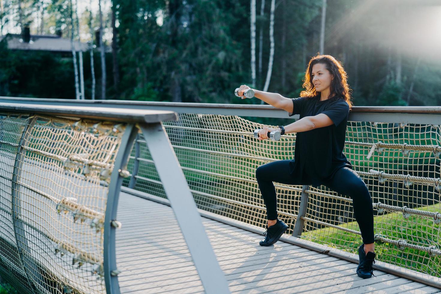 motivierte brünette aktive junge frau macht hockübungen mit hanteln, trainiert bizeps, in schwarzer aktiver kleidung gekleidet, posiert bei sonnenaufgang auf der brücke im freien, hat morgens training. Sportkonzept foto