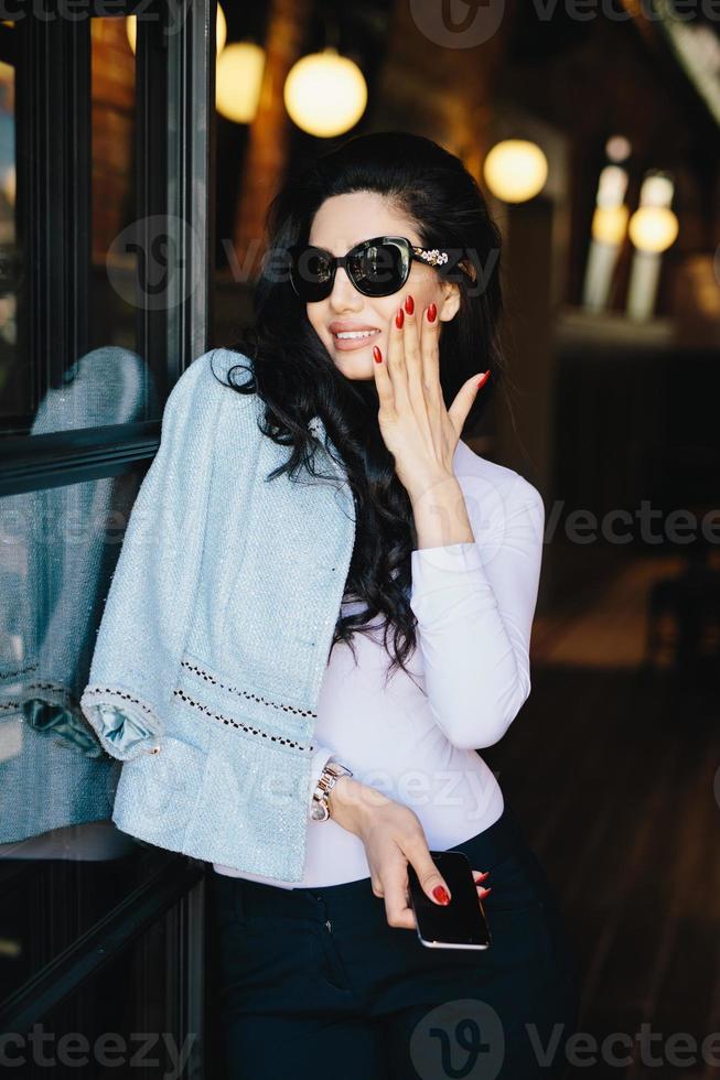 Vertikales Porträt einer glamourösen Geschäftsfrau mit dunklem, luxuriösem Haar, die eine Sonnenbrille und formelle Kleidung trägt und die Hand auf die Wange hält und ihre roten schönen Nägel mit dem Handy demonstriert, während sie im Café steht foto