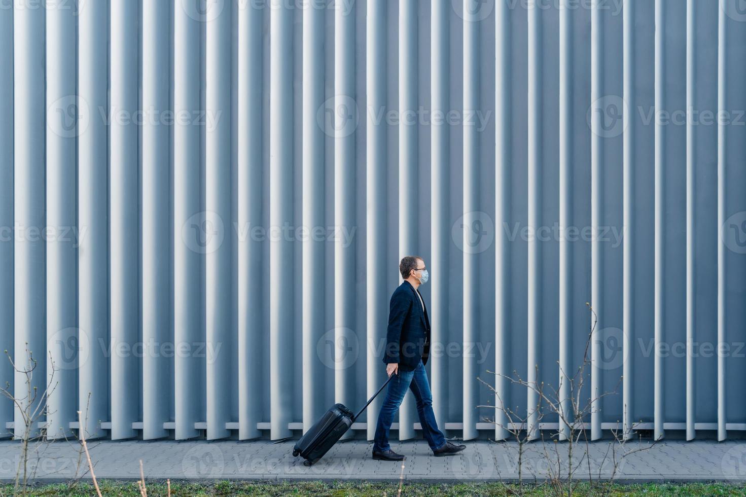 Horizontale Panoramaaufnahme eines Passagiers, der wegen der Quarantäne- und Pandemiesituation in der Welt im eigenen Land ankommt, mit Koffer geht, im Freien gegen Metallzaun posiert, Gesichtsmaske trägt foto