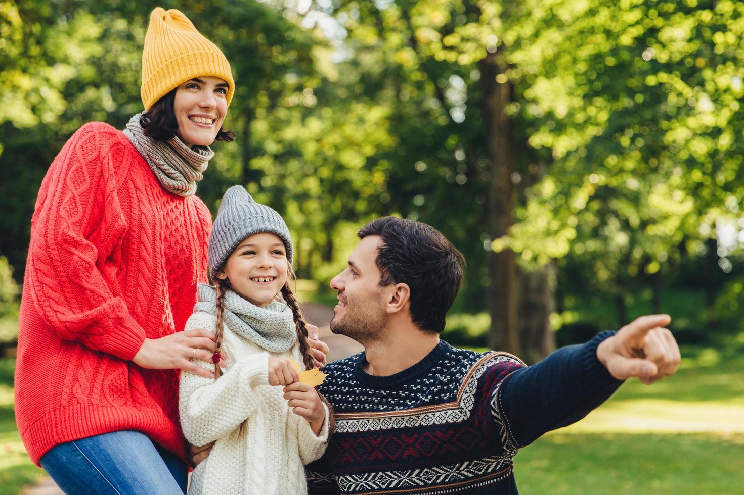 hübsches kleines mädchen mit zwei zöpfen, trägt strickmütze, schal und pullover, hält blatt in der hand, schaut freudig in die ferne, während ihr vater ihre schöne blume zeigt. Herbsttage zusammen verbringen foto