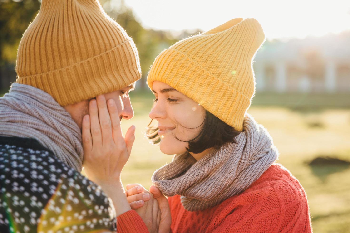 horizontales porträt einer brünetten frau mit warmem hut, schal und pullover, hält die hände auf der wange des freundes, hat einen romantischen moment und wird sich als dating küssen. Verliebte Paare sehen sich leidenschaftlich an foto