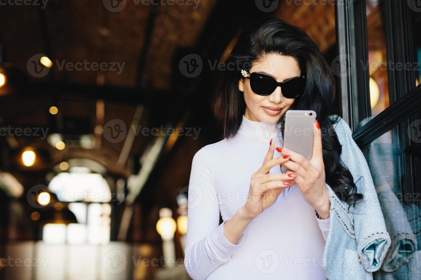 elegante brünette frau mit reiner haut, sonnenbrille tragend, mit roter perfekter maniküre in weißer formeller kleidung, handy mit kostenlosem internet, checken ihrer e-mails. Menschen, Kommunikation foto