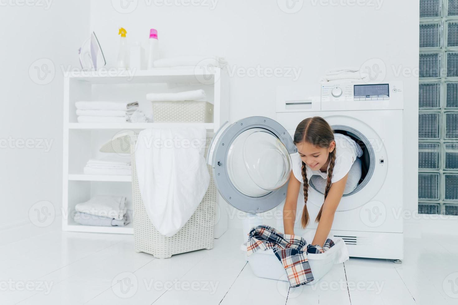 hübsche kleine Haushälterin posiert in Waschmaschine, nimmt kariertes Hemd aus Becken, wäscht Wäsche, hat fröhlichen Ausdruck, zwei gekämmte Zöpfe. Konzept für Kindheit und Waschtag foto